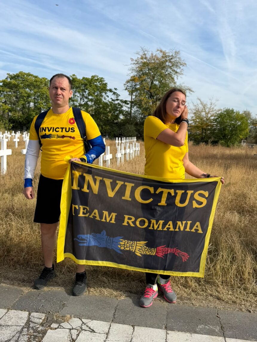 Two people wearing yellow Invictus shirts stand in a grassy area, holding a black and yellow Invictus Team Romania banner in front of a cemetery with white crosses.
