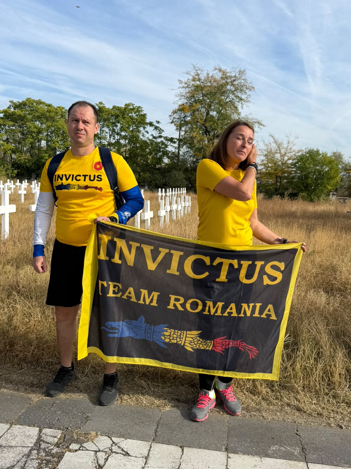 Two people wearing yellow Invictus shirts stand in a grassy area, holding a black and yellow Invictus Team Romania banner in front of a cemetery with white crosses.