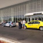Airport curb scene with a yellow taxi and a light blue hatchback as police speak with a man outside the departures entrance.