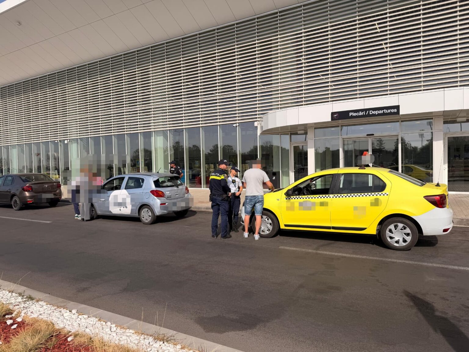 Airport curb scene with a yellow taxi and a light blue hatchback as police speak with a man outside the departures entrance.
