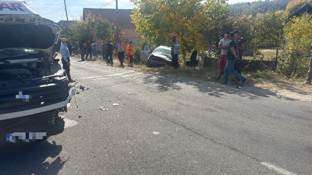 Front view of a white van with major front-end damage after a collision, on a rural road with onlookers nearby