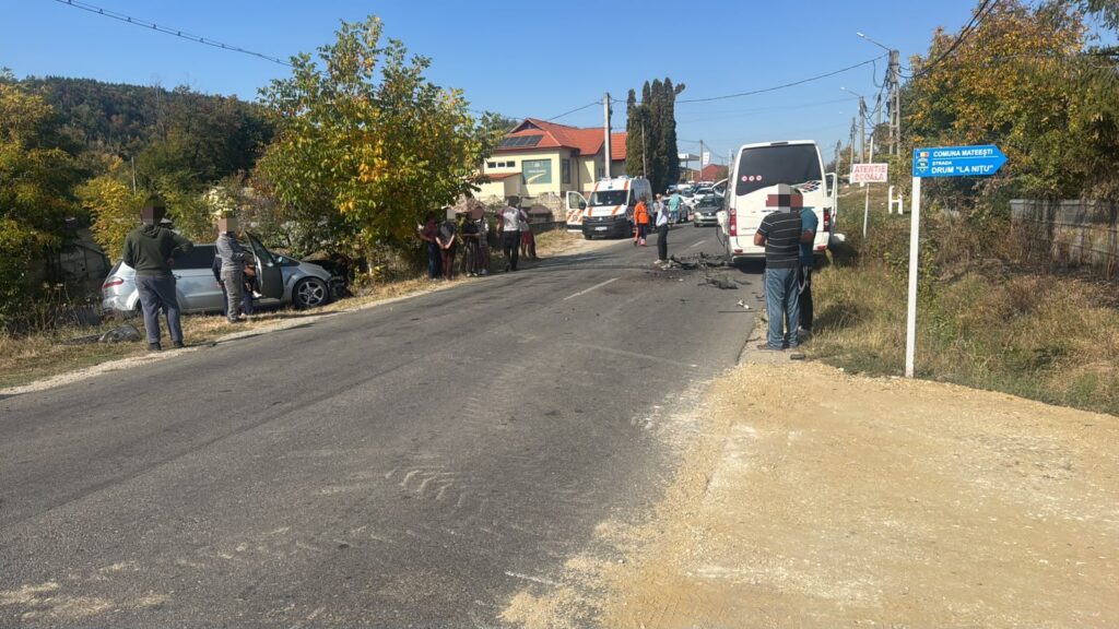 White car hit a tree on the roadside with bystanders and responders nearby, an ambulance behind, and a bus farther down the street on a rural road Hazard scene.