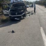 White delivery van heavily damaged at the front, hood open, debris scattered on a rural road, onlookers nearby.