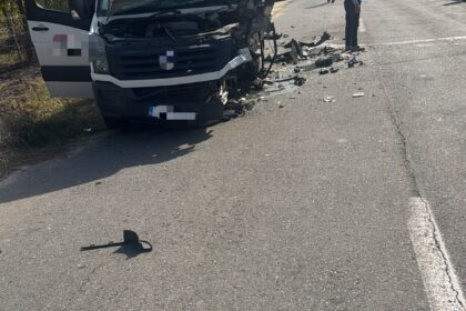 White delivery van heavily damaged at the front, hood open, debris scattered on a rural road, onlookers nearby.