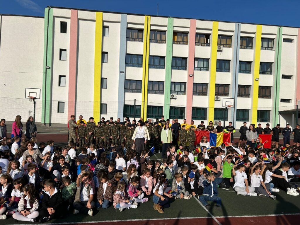 WhatsApp Image 2026-10-09 at 124217 - Jurnalul Olteniei – Știri din Craiova și Oltenia Large group of students gathered on a school court in front of a colorful multi-story building with vertical pastel stripes; some people in uniforms and several flags nearby.