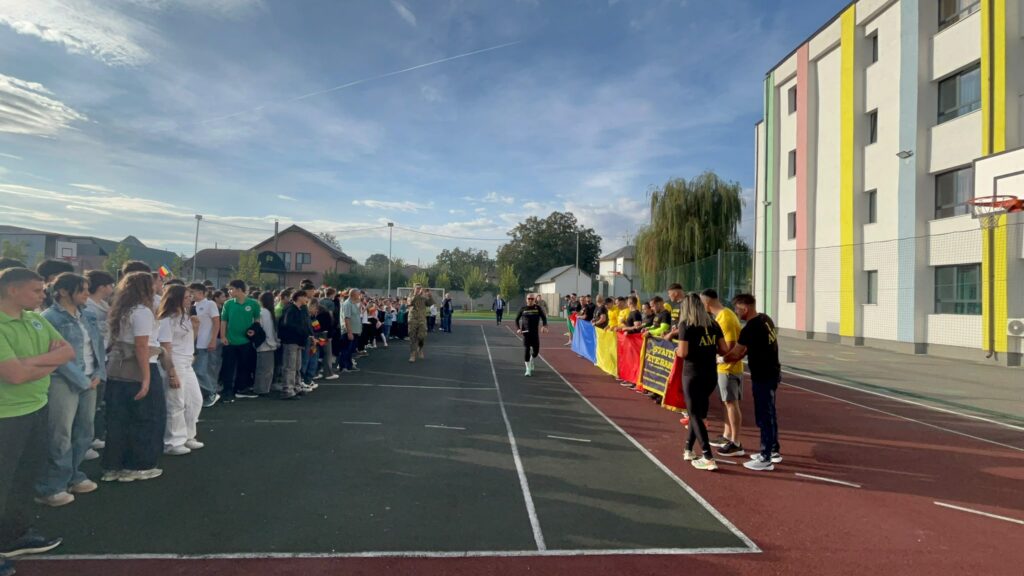 WhatsApp Image 2026-10-09 at 124217 11 - Jurnalul Olteniei – Știri din Craiova și Oltenia Line of students standing along a school track as a soldier walks toward them, with a large Romanian flag banner nearby and colorful building on the right.