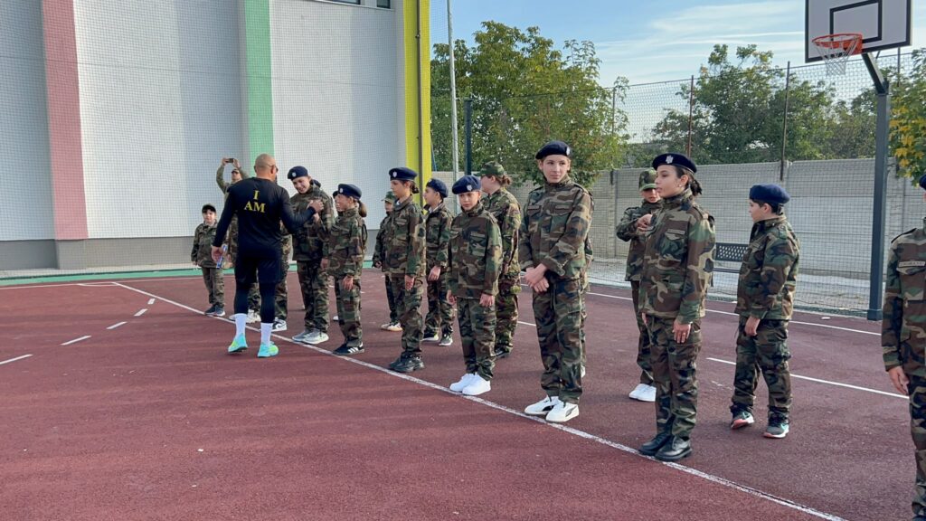WhatsApp Image 2026-10-09 at 124217 5 - Jurnalul Olteniei – Știri din Craiova și Oltenia Group of soldiers in camouflage uniforms lined up on an outdoor basketball court, listening to a trainer with'I AM' on his shirt.