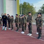 Group of soldiers in camouflage uniforms lined up on an outdoor basketball court, listening to a trainer with 'I AM' on his shirt.
