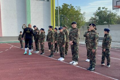Group of soldiers in camouflage uniforms lined up on an outdoor basketball court, listening to a trainer with 'I AM' on his shirt.
