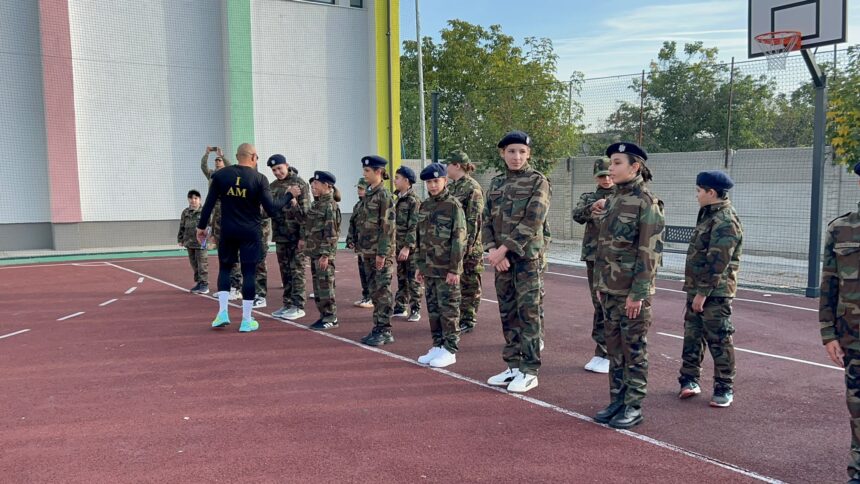 Group of soldiers in camouflage uniforms lined up on an outdoor basketball court, listening to a trainer with 'I AM' on his shirt.