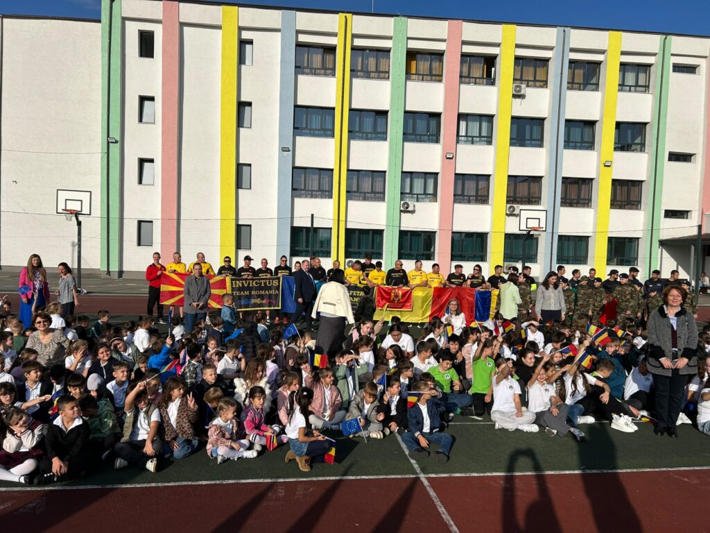WhatsApp Image 2026-10-09 at 124218 7 - Jurnalul Olteniei – Știri din Craiova și Oltenia Group of schoolchildren seated on a sports court, holding Romanian flags, with a colorful multi‑storey school building in the background.