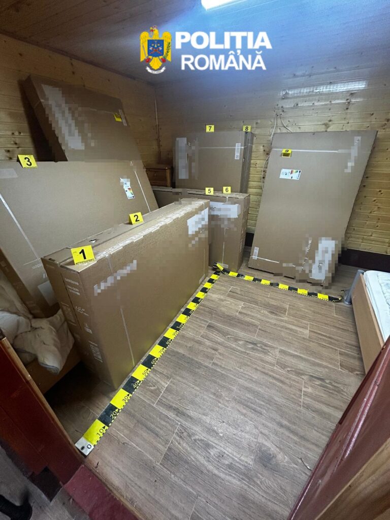 Small storage room with large cardboard boxes and appliances stacked along wooden walls, yellow numbered evidence markers on items, police watermark at top (Poliția Română).