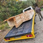 Romanian police crest above a man loading long wooden planks onto a yellow trailer on a forest road.