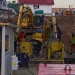 Construction site with yellow excavators digging amid piles of dirt, residential buildings in the background.