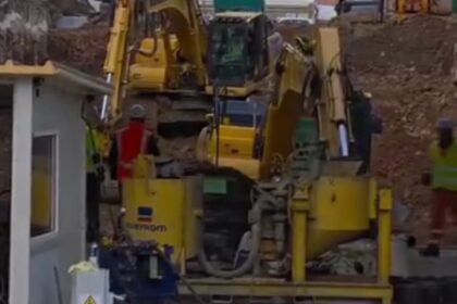Construction site with yellow excavators digging amid piles of dirt, residential buildings in the background.