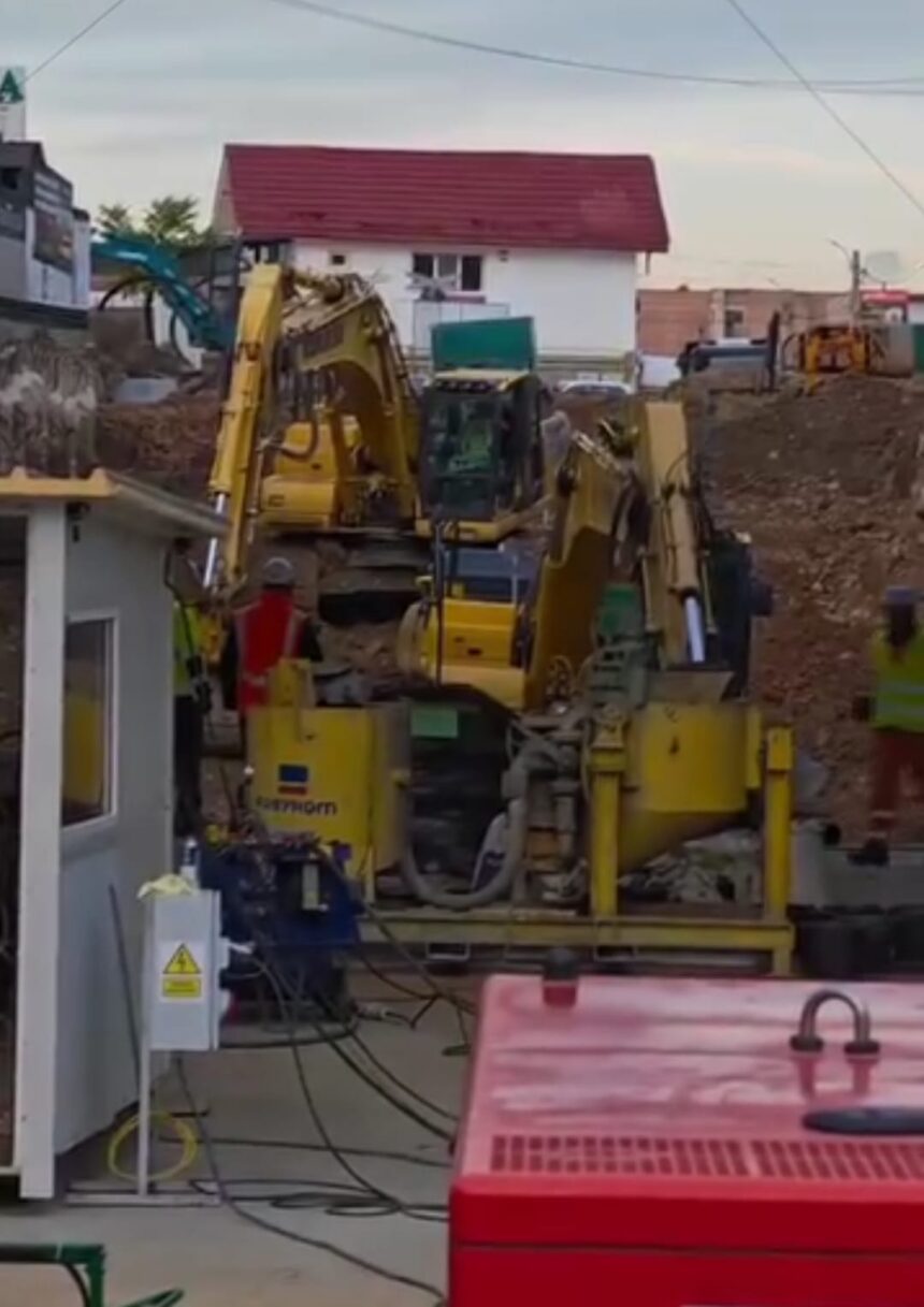 Construction site with yellow excavators digging amid piles of dirt, residential buildings in the background.