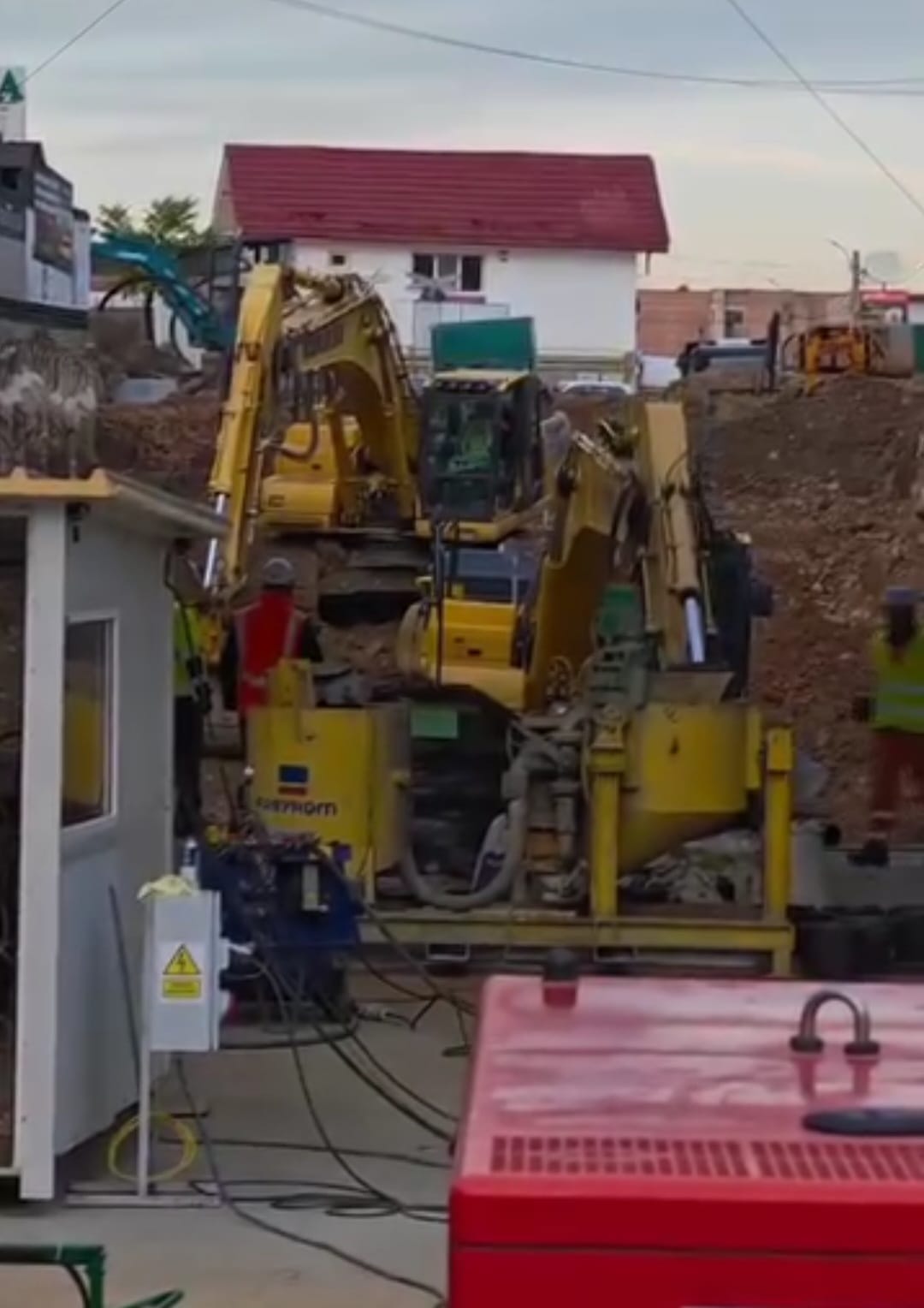 Construction site with yellow excavators digging amid piles of dirt, residential buildings in the background.