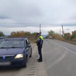 Police officer in a high-visibility vest chats with the driver of a blue sedan stopped on the roadside near a railroad crossing in a rural area.