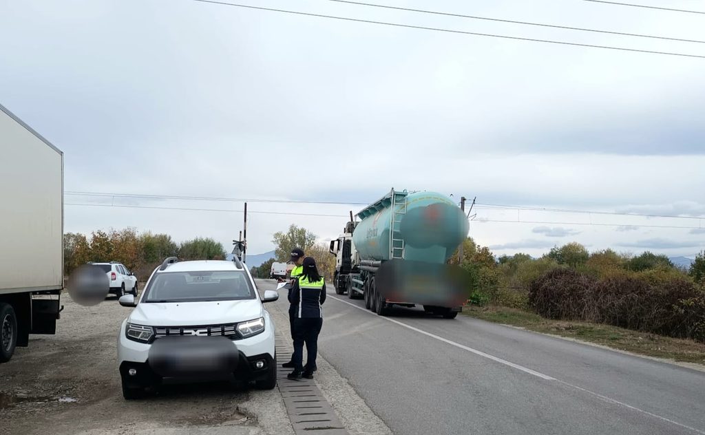 Two officials in hi-vis vests stand by a white SUV on the road shoulder as a tanker truck passes nearby.