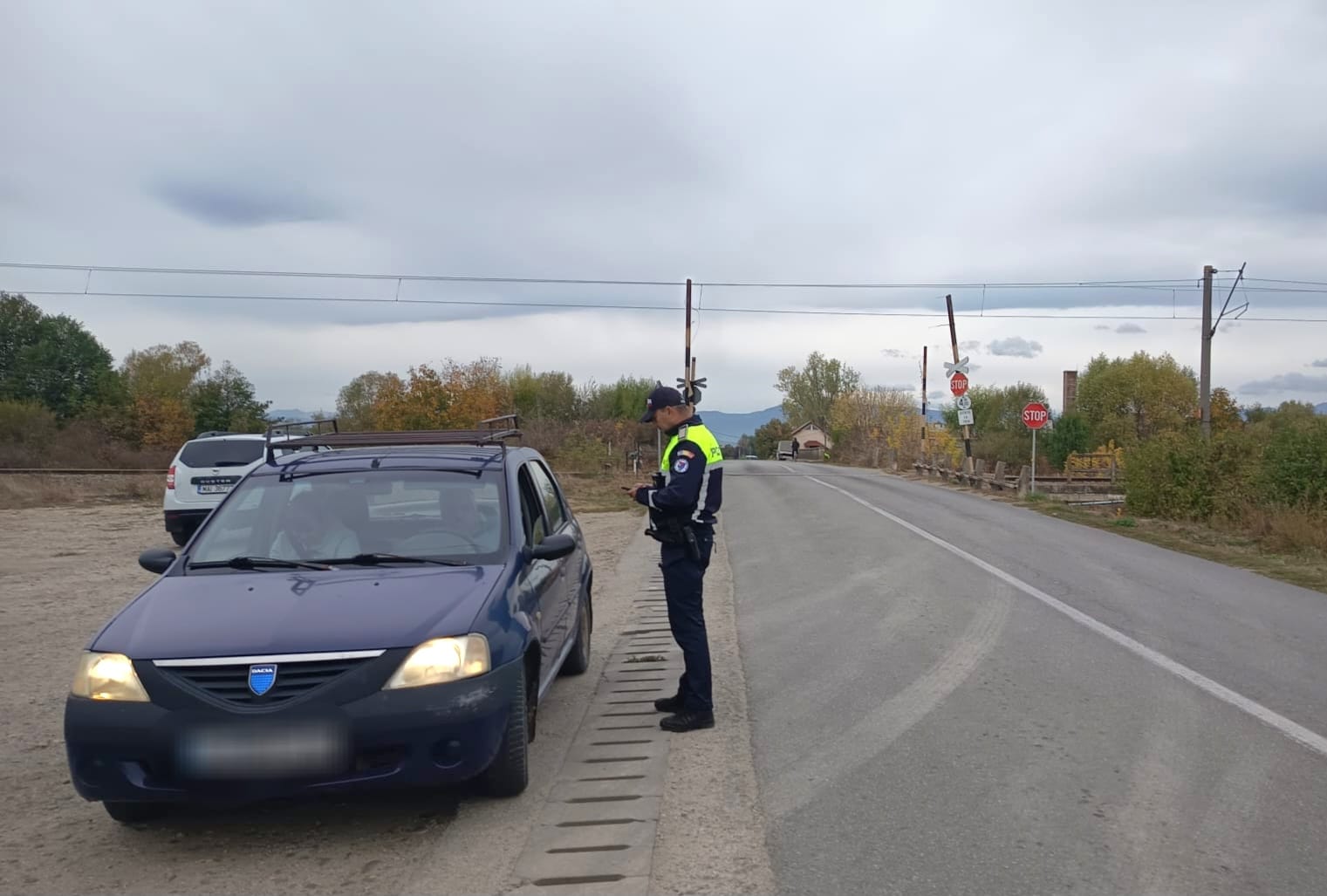 Police officer in a high-visibility vest chats with the driver of a blue sedan stopped on the roadside near a railroad crossing in a rural area.