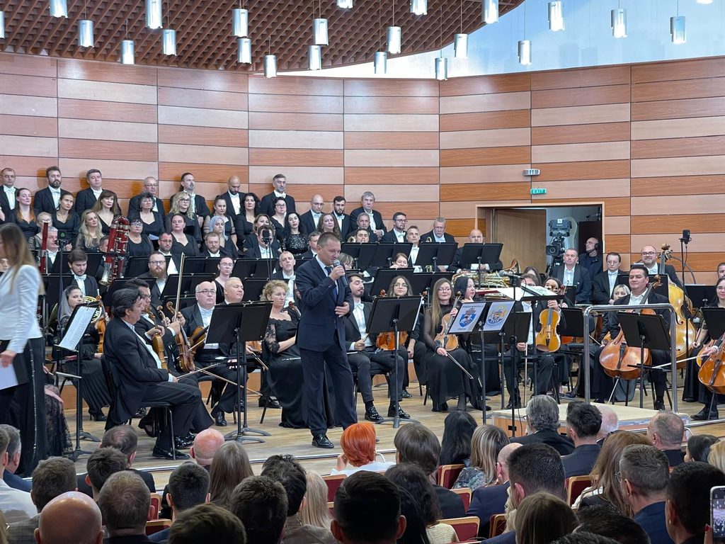 WhatsApp Image 2026-10-20 at 153635 - Jurnalul Olteniei – Știri din Craiova și Oltenia Conductor in a dark suit leads a large orchestra and choir during a formal concert in a wood-paneled hall.