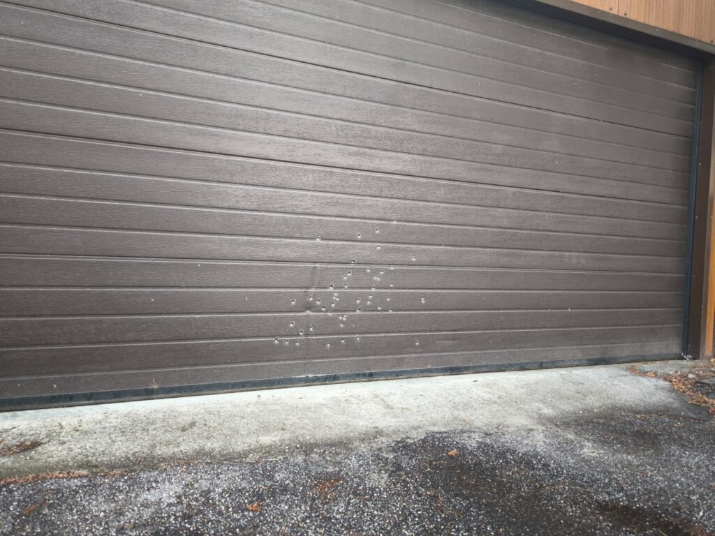WhatsApp Image 2026-10-22 at 152114 - Jurnalul Olteniei – Știri din Craiova și Oltenia Brown horizontal-paneled garage door with a cluster of small white water droplets toward the bottom center.