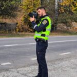 Police officer in a neon vest uses a handheld camera on a roadside with autumn trees in the background.