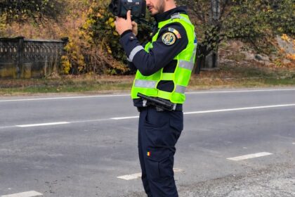 Police officer in a neon vest uses a handheld camera on a roadside with autumn trees in the background.