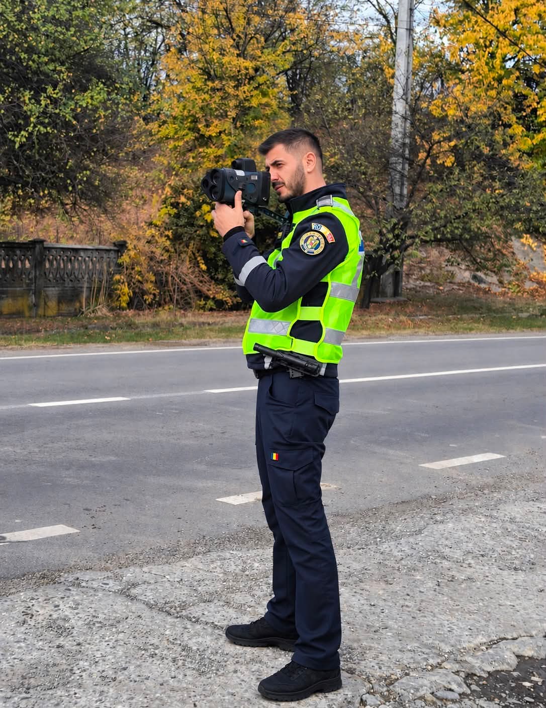 WhatsApp Image 2026-10-29 at 121606 - Jurnalul Olteniei – Știri din Craiova și Oltenia Police officer in a neon vest uses a handheld camera on a roadside with autumn trees in the background.