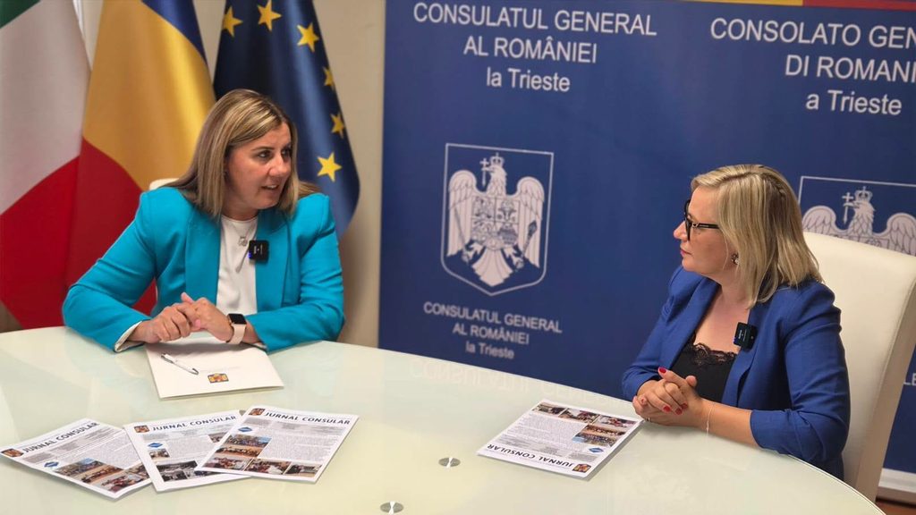 WhatsApp Image 2026-10-29 at 140310 - Jurnalul Olteniei – Știri din Craiova și Oltenia Two women in blue blazers converse at a glass table in a Romanian consulate office, with flags and a 'Consulatul General al României' backdrop behind them.
