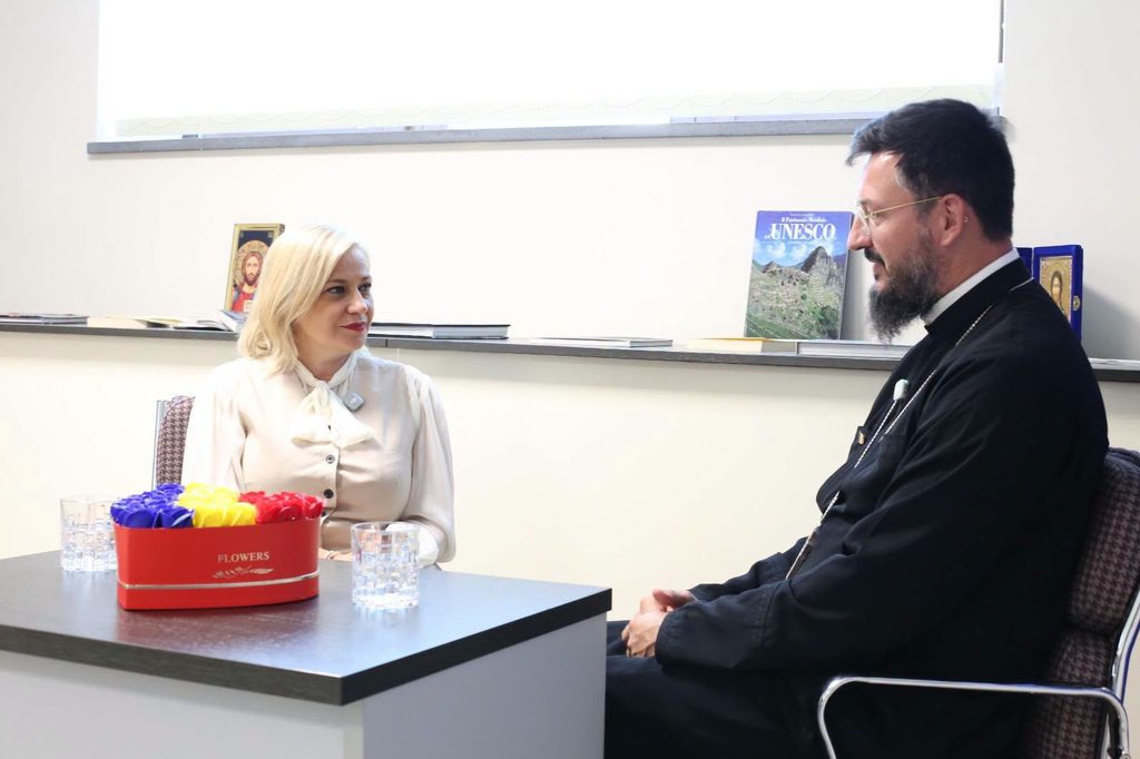 WhatsApp Image 2026-10-29 at 140311 - Jurnalul Olteniei – Știri din Craiova și Oltenia Woman with blonde hair sits across from a bearded man in clerical clothing at a desk; a red box of colorful flowers sits foregrounded on the table.