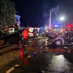Nighttime emergency scene with firefighters inspecting a multi-vehicle crash; rain, debris, and a fire truck with flashing lights in the background.