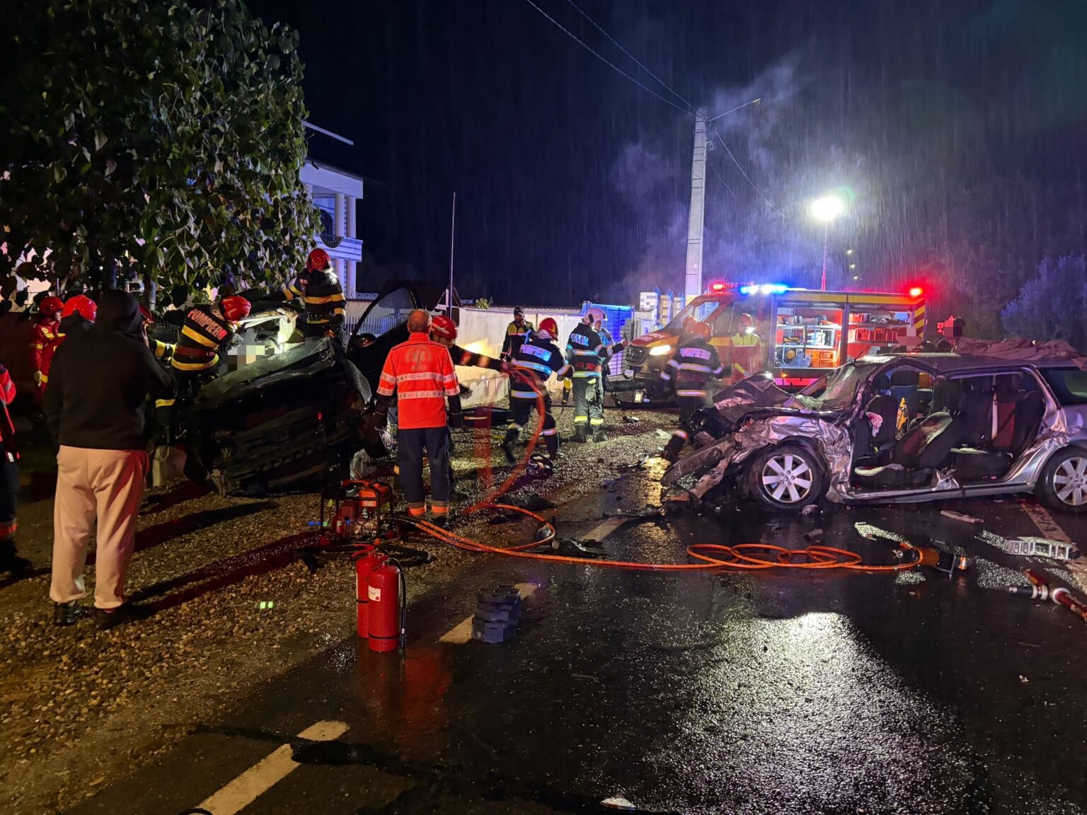 Nighttime emergency scene with firefighters inspecting a multi-vehicle crash; rain, debris, and a fire truck with flashing lights in the background.