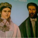 Portrait of a woman in an ornate headdress and white gown beside a bearded man in a dark suit against a blue background.