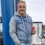 Man in a denim jacket stands at a gas station, smiling and holding a red cup, with a fuel pump in the background.