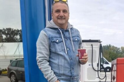 Man in a denim jacket stands at a gas station, smiling and holding a red cup, with a fuel pump in the background.