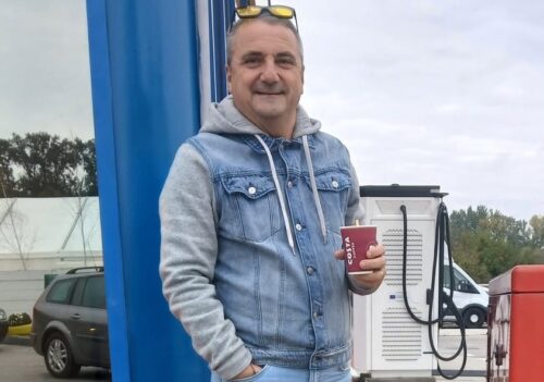 andronesi-e1792438845192 - Jurnalul Olteniei – Știri din Craiova și Oltenia Man in a denim jacket stands at a gas station, smiling and holding a red cup, with a fuel pump in the background.