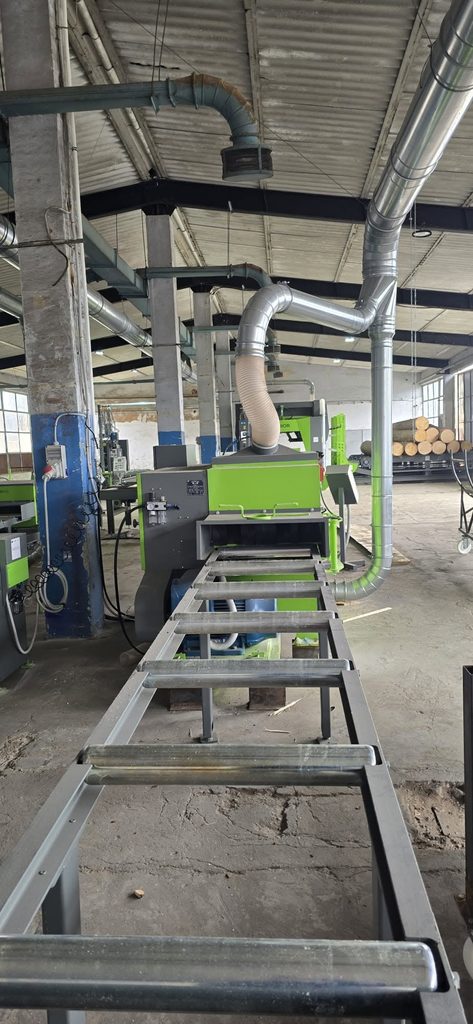 Industrial workshop with green machinery and large silver dust-collection ducts; metal framework and logs visible in the background.
