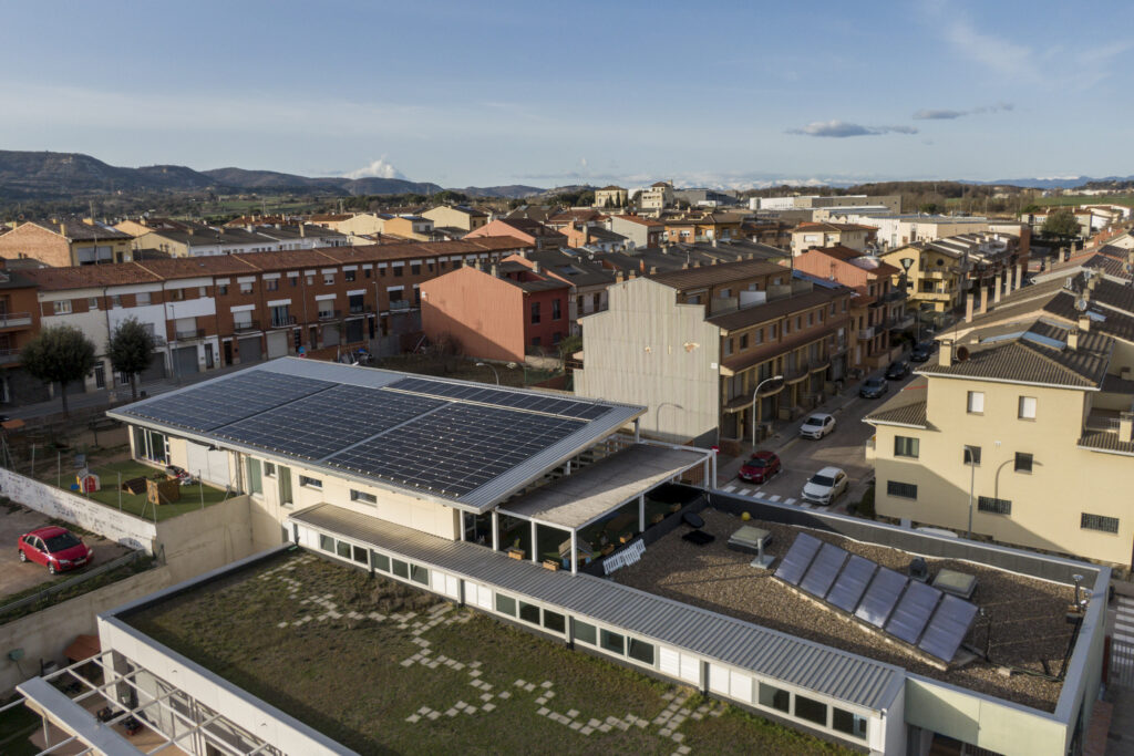 Aerial view of a town with red-tiled roofs and a large flat building covered in solar panels in the foreground, hills in the distance.