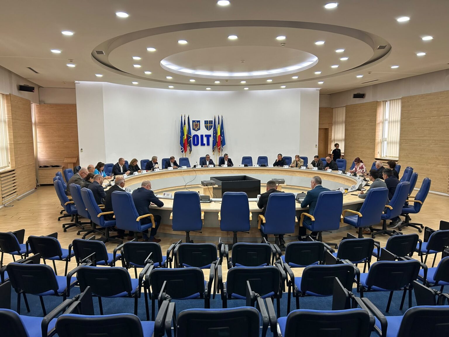 consiliu judetean cj olt 2026-10-15 at 132325 - Jurnalul Olteniei – Știri din Craiova și Oltenia Large conference room with a circular wooden table and many attendees seated in blue chairs, participating in a formal meeting; flags and a crest are displayed at the far wall behind the speakers.