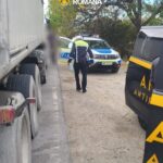 Romanian police officer inspecting a stopped truck on a gravel road with a police car in the background.