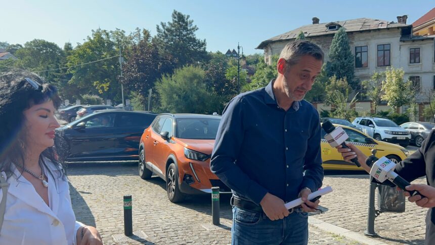 Man in a blue shirt holds papers and speaks into multiple microphones during an outdoor interview; a woman in a white shirt stands nearby on a cobblestone street with parked cars and trees in the background.