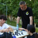 Group of people playing chess at an outdoor park table, with one young man wearing white t-shirt and earbud looking at the board while others watch nearby.