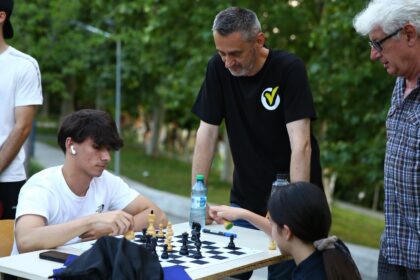 Group of people playing chess at an outdoor park table, with one young man wearing white t-shirt and earbud looking at the board while others watch nearby.