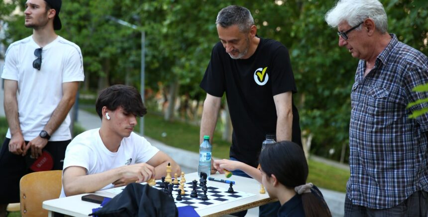 Group of people playing chess at an outdoor park table, with one young man wearing white t-shirt and earbud looking at the board while others watch nearby.