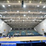 Indoor basketball arena with players in blue jerseys warming up on a polished court; large, tiered blue and white seats surround the court and banners hang along the walls.