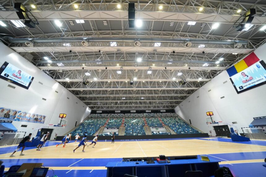 Indoor basketball arena with players in blue jerseys warming up on a polished court; large, tiered blue and white seats surround the court and banners hang along the walls.