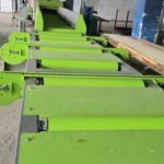 Green industrial conveyor system with rollers and guides in a dusty workshop, with overhead ducts and machinery in the background.