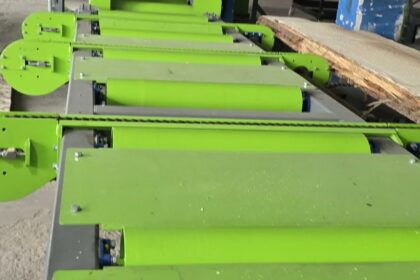 Green industrial conveyor system with rollers and guides in a dusty workshop, with overhead ducts and machinery in the background.
