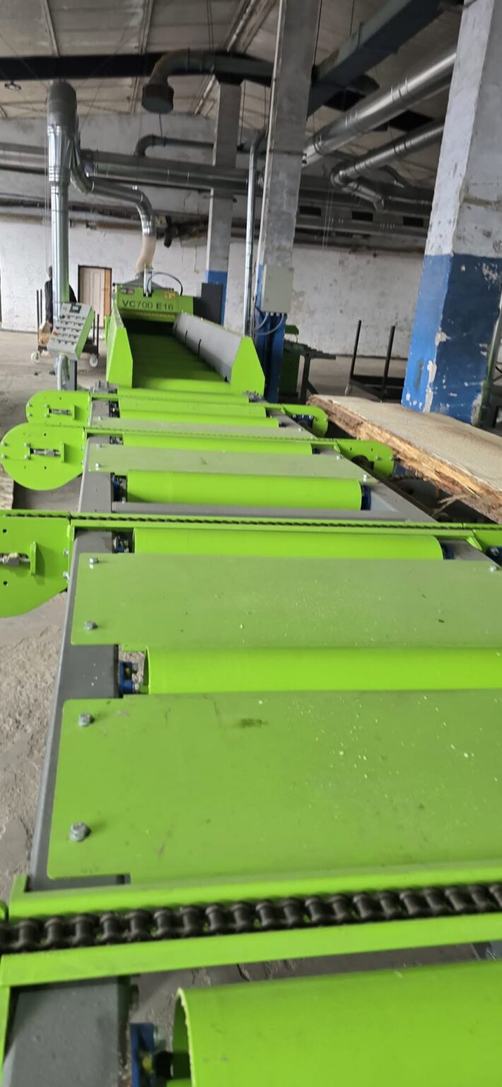 Green industrial conveyor system with rollers and guides in a dusty workshop, with overhead ducts and machinery in the background.